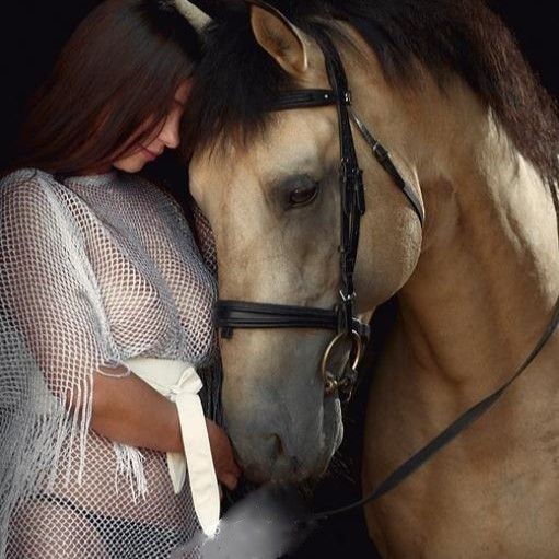 Woman in sheer white outfit standing closely with a brown horse, both looking downwards.